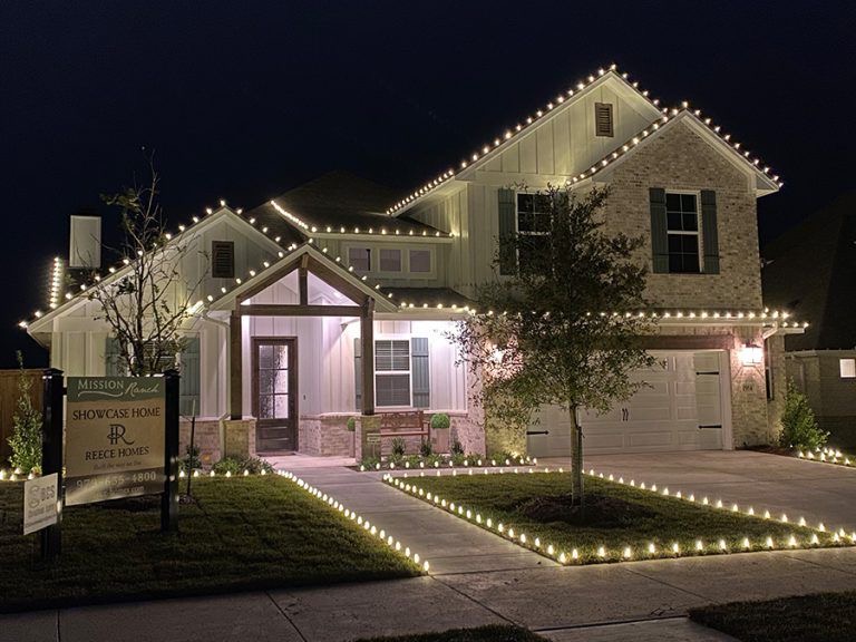 White two-story farmhouse with warm white roofline lights and lit walkway stakes